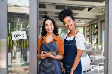  Two women stand smiling in the doorway of a restaurant or cafe. The space behind them is filled with square wooden tables and white chairs; baskets with plants growing out of them are mounted on the walls. The woman on the left wears an orange collared shirt under a dark-blue-and-white-striped apron. She holds a digital tablet. To the left of her is the open door, which has an open sign hanging from a suction cup on its window. The woman on the right is slightly taller and wears a white T-shirt under a dark blue apron. 
