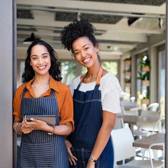  Two women stand smiling in the doorway of a restaurant or cafe. The space behind them is filled with square wooden tables and white chairs; baskets with plants growing out of them are mounted on the walls. The woman on the left wears an orange collared shirt under a dark-blue-and-white-striped apron. She holds a digital tablet. To the left of her is the open door, which has an open sign hanging from a suction cup on its window. The woman on the right is slightly taller and wears a white T-shirt under a dark blue apron. 