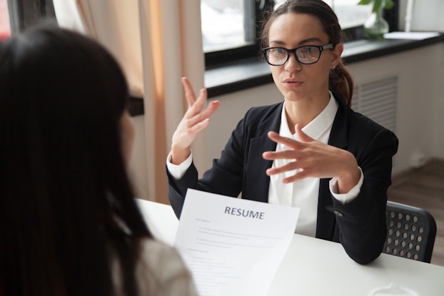 A woman in glasses talks to another woman, who faces away from the camera and holds the first woman's resume.