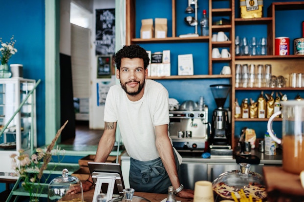  A young man with dark hair and a beard leans over a counter in a cafe. The wall behind him is painted a deep blue and filled with shelving holding cups, mugs, and bags and cans of coffee. The stainless steel counter below the shelves holds two coffee machines, another bag of coffee and a carton of creamer. The front counter that the man leans on is filled with plates of pastries covers with glass lids and an electronic tablet used as a point of service. The man wears a white T-shirt and dark gray apron. He has a slight smile on his face.
