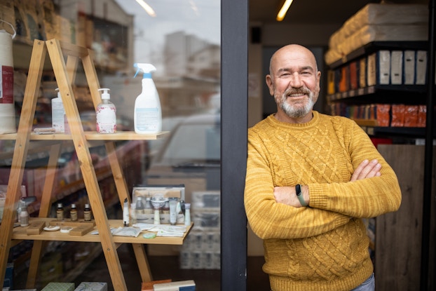  A man in a yellow sweater stands with his arms crossed in the doorway of a shop. The man is bald with a white beard and he leans against the doorjamb with a confident grin on his face. Beside the entrances is a wooden shelving unit that resembles a ladder with the rungs replaced with planks of wood, which are used to display cleaning products, bottles of hand soap, and small vials of perfume oils.