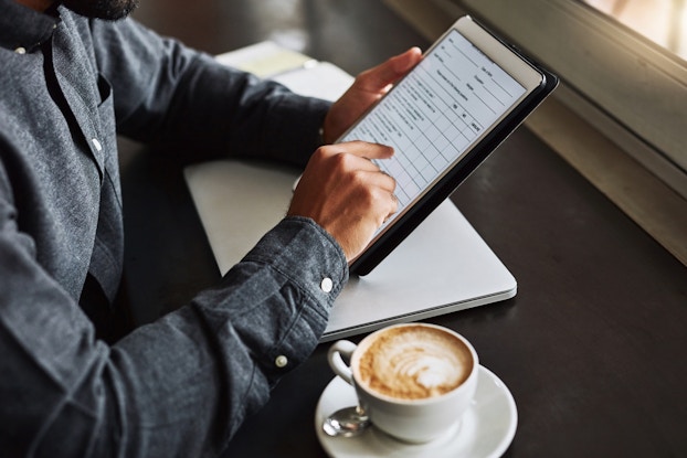  A below-the-shoulders shot of a man sitting at a dark wood table, using a tablet to fill out a form. A coffee cup and saucer sits to his right.