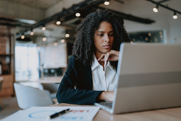 A young woman sits at a table, working at a laptop with a thoughtful look on her face. She wears a white collared shirt and a black blazer. A chart and a pen sit next to the laptop on the table.