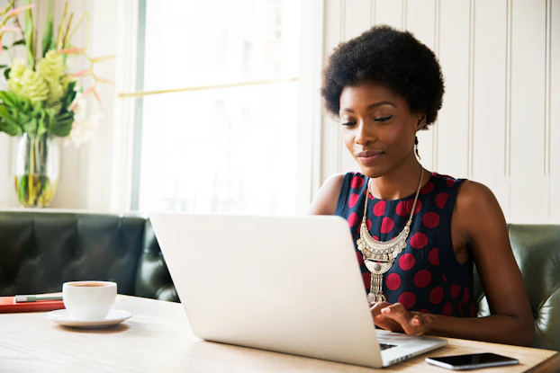 A woman is sitting in a black leather booth at a table, typing on a laptop. The woman has Afro hair, and she is wearing a navy blue sleeveless blouse with red polka dots and a necklace with small gold coins dangling from it.