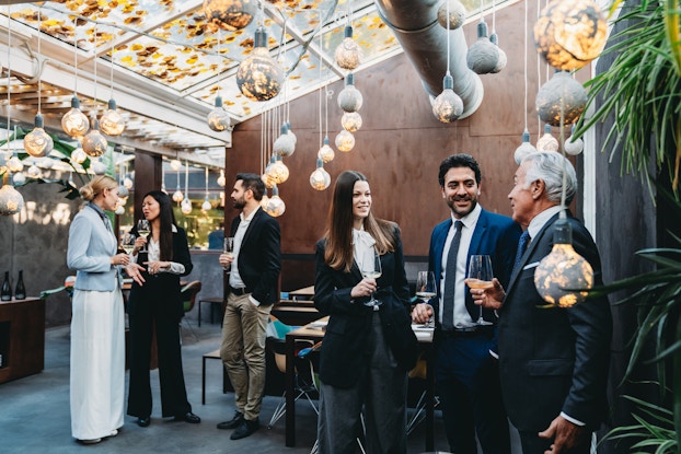 Six people in businesswear stand in groups of three and mingle with wine glasses in their hands at an indoor event. Their meeting space has gray carpeting and brown walls textured to look like wood. The ceiling is slanted and made of glass; yellow leaves have fallen onto it and partially obscure the view of the sky. Painted lightbulbs hang down from the ceiling, some of them low enough to be near a person's head or shoulder. The bulbs have varying amounts of gray swirling paint on them -- some bulbs have light shining through the paint, but others are completely covered.