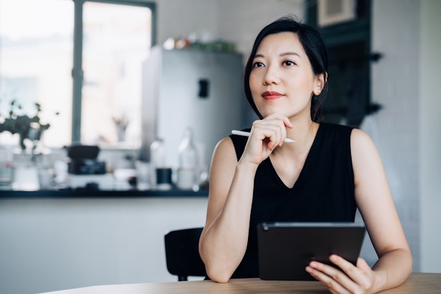 A young woman sits at a table and looks into the distance in deep thought. One of the woman's hands holds a digital tablet and the other hand is held to her chin. The woman has black hair pulled back, and she wears a black sleeveless top.
