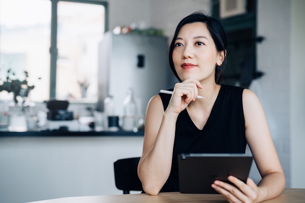  A young woman sits at a table and looks into the distance in deep thought. One of the woman's hands holds a digital tablet and the other hand is held to her chin. The woman has black hair pulled back, and she wears a black sleeveless top.