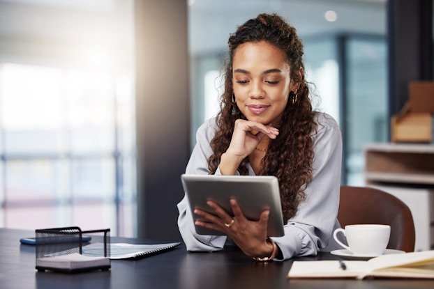 A young woman sits at a table and looks at the electronic tablet that she holds in one hand. Her other hand is on her chin, and she is smiling. A coffee cup and an open book sit next to her on one side of the table; some papers and a smartphone sit on the other side.