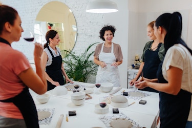  A group of women in aprons stand around a table in a large white room. Each woman has a circle of gray clay sitting on a placemat in front of her on the table; the table also holds several white ceramic bowls, a few carving tools, and enough glasses of red wine for everyone. The woman closest to the camera, standing in the foreground and facing away from the viewer, is taking a sip of wine. Most of the women are wearing black aprons, but the woman in the center of the image is wearing a white one. The other women are turned to face the woman in the white apron, who is smiling at the group. 