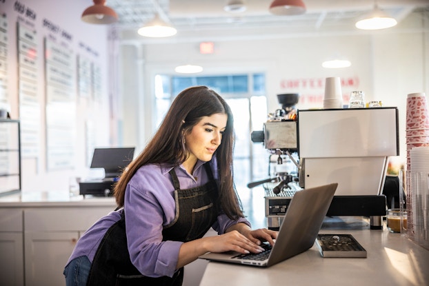 A woman stands behind the counter at a cafe, typing on a laptop. The woman has long brown hair and she wears a dark denim apron over a lavender button-up shirt and jeans. The cafe is all white, with the shop's offerings listed on signs along one of the walls. The L-shaped counter holds a large coffee machines and stacks of paper cups.