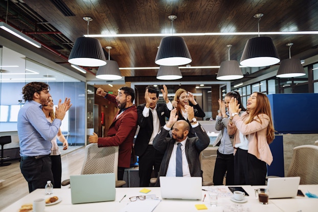 A group of eight people in business casual attire sit and stand around one side of a conference table in a large open office space. The people have smiles on their faces and their hands in the air, applauding and celebrating.