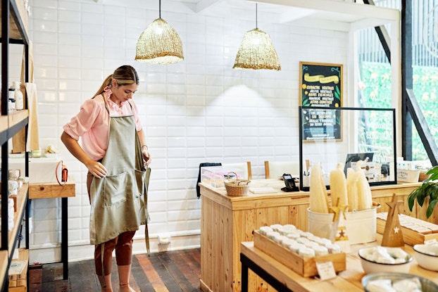 The owner of a bath and body retail store stands behind the counter and puts on an apron. She is preparing as she opens her store for the day.