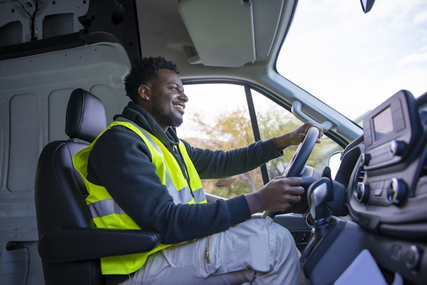  A profile shot of a man in the driver's seat, taken in the interior of a delivery van. The man is young and wearing gray cargo pants, a long-sleeved dark blue shirt, and a yellow high-visibility vest. He has both hands on the steering wheel and is smiling while looking out through the windshield.