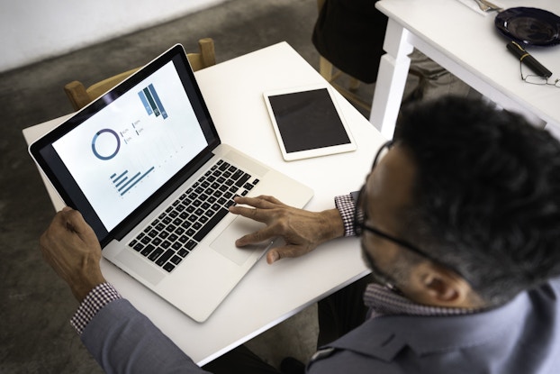 A man, seen from above, looks at a series of charts on a laptop screen. On the table next to his laptop is a smart tablet.