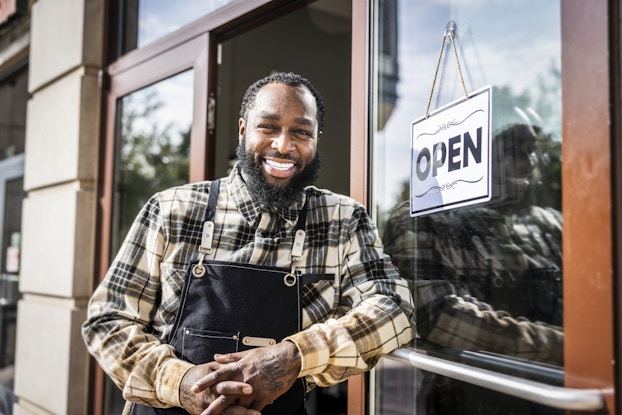 A man stands outside of his business, leaning against an open door hung with an "OPEN" sign. The man has short black hair and a black beard. He is smiling and wearing a light yellow plaid shirt and a dark blue apron.