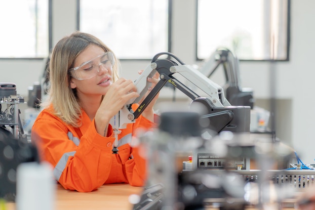  A woman in a bright orange jumpsuit uses her hands to adjust the end of a small robotic arm. The woman has shoulder-length blonde hair and is wearing goggles to protect her eyes.
