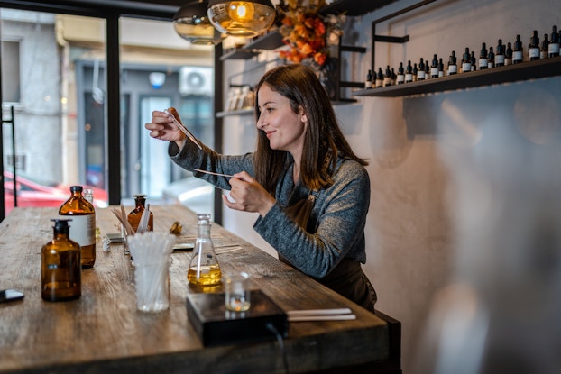 A woman stands behind a worn wooden counter in a perfumery, sampling a scent. She holds an eye dropper over a thin strip of paper, preparing to drop the fragrance oil onto the strip. The woman has shoulder-length brown hair and she wears a long-sleeved gray top. On the counter in front of her are several bottles of liquid, as well as more paper strips and a jar of eye droppers. On a shelf behind her are a few dozen small brown bottles with eye dropper tops.