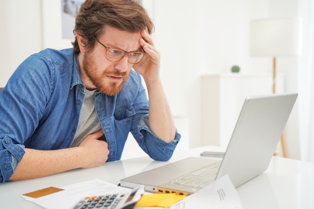 A man looks at his laptop with one hand on his head. His face is worried. Beside the laptop are some papers and a calculator.
