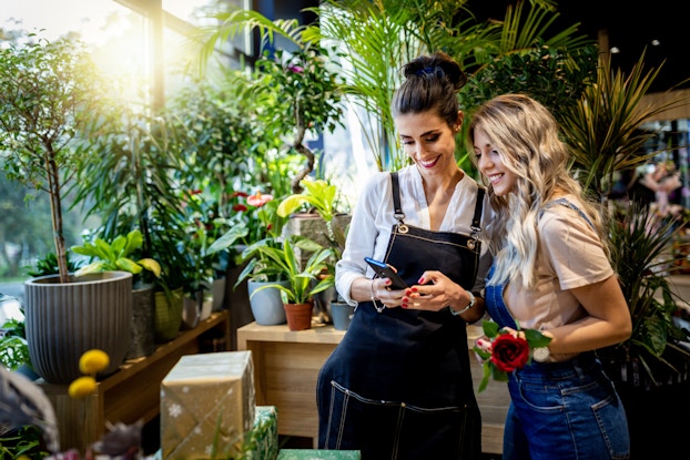 Two smiling women in denim aprons stand in a plant shop and look at something on a smartphone. The woman on the left is holding the smartphone; she has dark hair pulled into a bun on top of her head. The woman on the right is slightly shorter and has long, wavy blonde hair. The women are surrounded by green, leafy potted plants, some of which sit on top of wooden boxes.