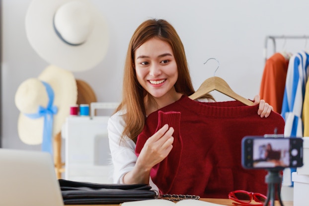 A woman holds up a red sweater on a hanger and looks in the direction of a smartphone on a tripod in the foreground. The smartphone is positioned horizontally and the image of the woman on the sweater can be seen out-of-focus on its touchscreen. In the background are more sweaters on hangers hanging on a rack and another rack holding wide-brimmed sunhats.