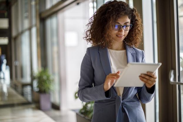 A young woman in a blazer stands in a hallway with floor-to-ceiling windows and examines an electronic tablet.