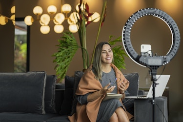  Woman sitting down, holding a notebook and recording herself with a ring light. 