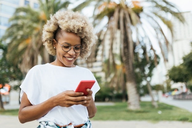  A woman with a curly blonde afro looks at the red smartphone in her hands with a smile. Out of focus in the background is a line of palm trees, with some buildings further off.