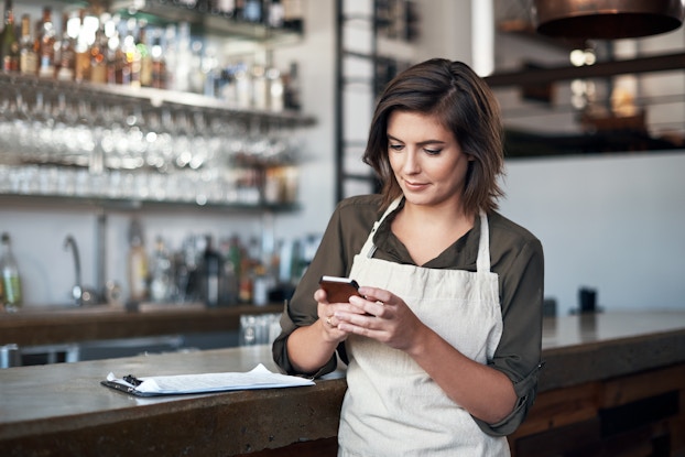  A woman stands with one elbow on the bar counter in a restaurant or bar. She looks down at the smartphone in her hand. She has shoulder-length brown hair and wears a cream-colored apron over an olive green collared shirt. A clipboard sits on the counter by her elbow. Behind the counter, out of focus, is another counter and several shelves filled with bottles and glasses.