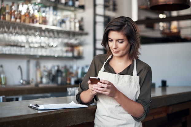 A woman stands with one elbow on the bar counter in a restaurant or bar. She looks down at the smartphone in her hand. She has shoulder-length brown hair and wears a cream-colored apron over an olive green collared shirt. A clipboard sits on the counter by her elbow. Behind the counter, out of focus, is another counter and several shelves filled with bottles and glasses.