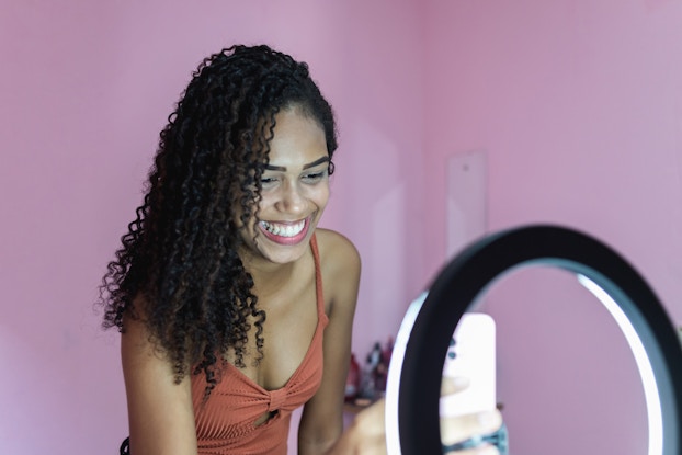  Woman standing in front of a pink background recording herself on her phone with a ring light.