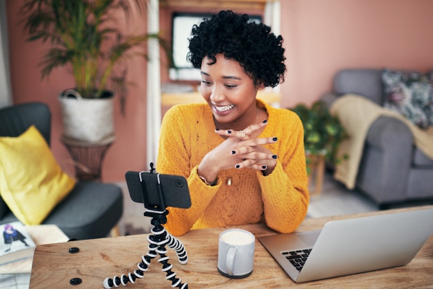  Smiling woman at home recording herself with her phone on a tripod.