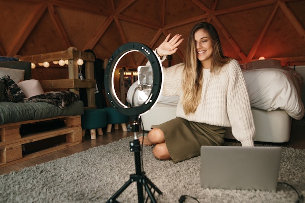  A young woman sits on a plush gray carpet and waves at a ring light attached to a tripod. She's sitting on the floor of a wood-panelled bedroom; behind her is a bed and beside her are a couple of small stools, a cushioned chair with a base made out of wooden pallets and a wooden railing wrapped in a string of lights.