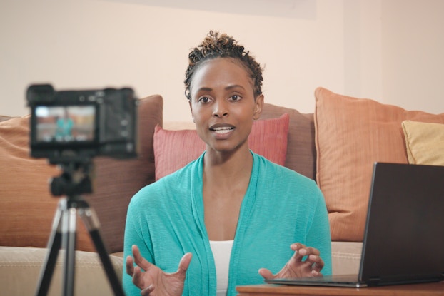  A woman in a teal cardigan sits in front of a sofa and speaks to a digital camera, which sits out of focus on a tripod in the foreground. An open laptop sits on a low table to the woman's left. The sofa behind the woman is piled with pillows in various sizes and shades of red and brown.