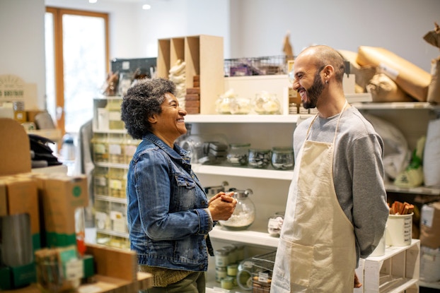 Two individuals, a man and woman, smile and laugh as they talk. The man is the owner of a store and is talking to the female customer.