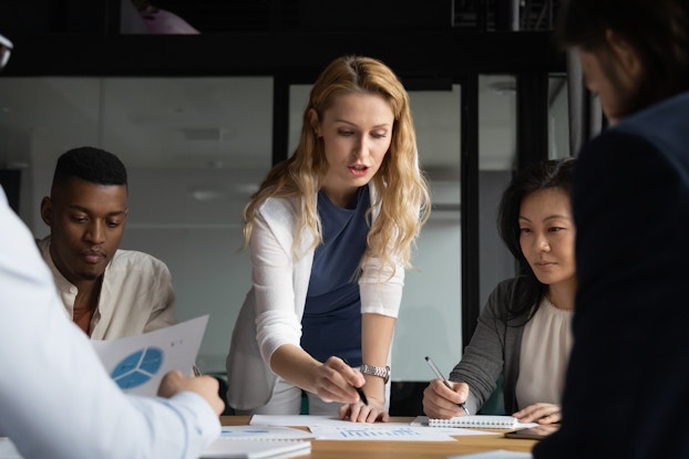 A diverse group of five people sits around a table for a meeting. One person, a blonde White woman, stands and looks down at a paper on the table, appearing to read from it. The others around the table consult their own papers.