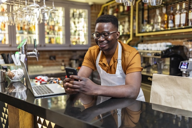  A man in an orange shirt and white apron leans on a glass-topped bar and looks at the smartphone in his hands. On the bartop beside him is an open laptop. In the background, shelves and glass-fronted cabinets are filled with liquor bottles.