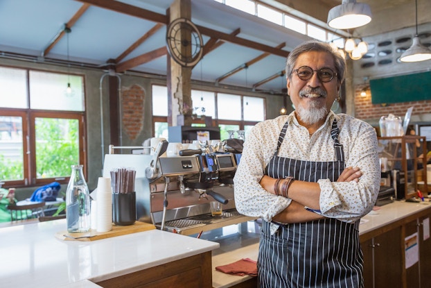 An older man in a striped apron stands in front of a coffee shop counter with his arms crossed and a proud smile on his face. The counter behind him is lined with stainless steel coffee machines, stacked cups and containers filled with straws.
