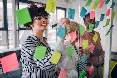  Three coworkers are seen through floor-to-ceiling glass panels; they are writing on the multi-colored post-it notes stuck to the glass. From left to right, the three coworkers are a curly-haired woman wearing glasses and a striped shirt; a shorter person with blond hair, their face obscured by the post-it notes; and a man wearing a salmon-colored button-up shirt. 