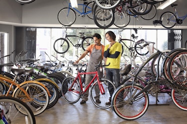 Two men stand in a large showroom filled with bicycles of various colors. The man on the left, who wears an orange shirt and gray apron, uses his hands to hold up a red bike. The man on the right, who wears a yellow T-shirt over a long-sleeved navy blue shirt, looks at the red bike with interests. Several more bikes hang from the ceiling of the showroom.