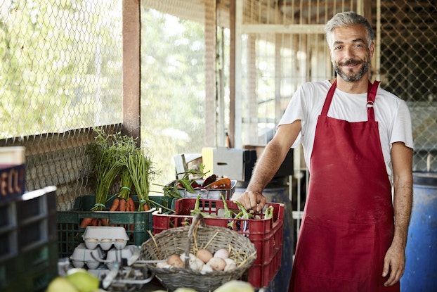 An older man with graying hair and a salt-and-pepper beard stands next to a table covered with crates and baskets of farm-harvested goods, including carrots, eggs, and eggplants. The man is wearing a red apron over a white T-shirt and he's smiling widely. In the background are makeshift walls made of chicken wire and wooden beams.
