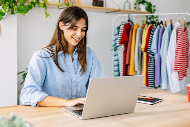 A smiling woman sits at a table and types on a laptop. The woman has long brown hair and is wearing a light blue button-up shirt. In the background is a clothing rack filled with hanging shirts, many of them with horizontal stripes.