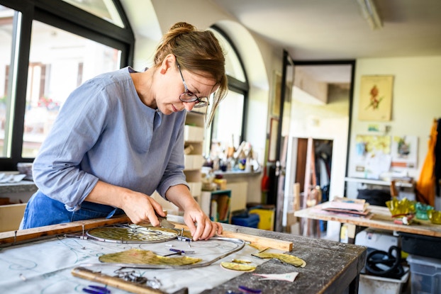 A glass artist leans over a table and carefully assembles the various pieces of a design she is working on.
