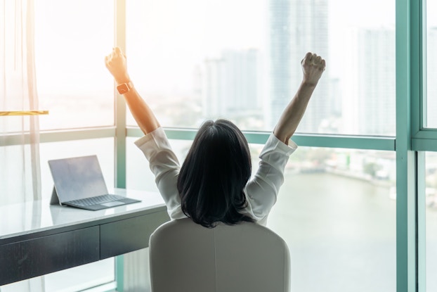 Woman sitting at desk with her back to the camera and her arms in the air with excitement.
