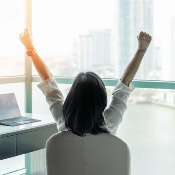 Woman sitting at desk with her back to the camera and her arms in the air with excitement.