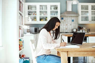 Woman sitting at kitchen table writing with pen and paper.