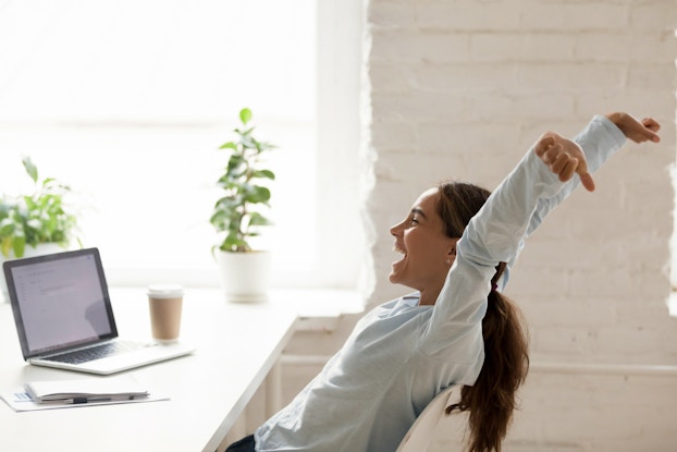A woman sitting at a desk leans back in her chair and stretches her arms over her head with a smile. A laptop sits open on the desk in front of her and two large plants sit on the sill of a large window in the background.