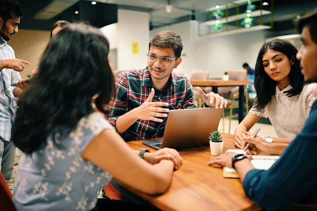 A group of young people in casual clothing sits around a table in a large open-concept office space. The camera's focus is on a young man in the middle, sitting in front of an open laptop and talking to the group while gesturing animatedly with his hands. He has brown hair and a mustache, and he wears glasses and a red-and-green plaid shirt.