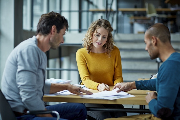 Three people sit around a table and examine a slim stack of papers. The man on the left, seen out of focus and in profile and wearing a light blue shirt, points at something on the paper. The woman in the middle, facing the viewer and wearing a yellow shirt, points at a different part of the page. The man on the right, also seen in profile and wearing a blue shirt, is lifting the page with one hand.