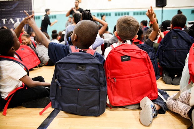  Young kids wearing State Bags backpacks sitting in a gymnasium.
