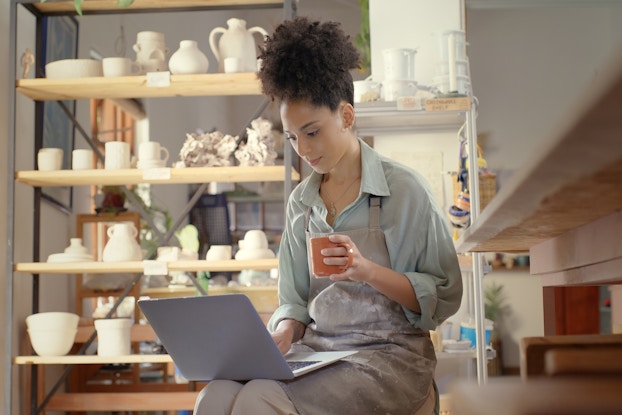 A woman sits in a workshop surrounded by shelves of uncoated ceramic cups and vases. With one hand, she types on the laptop perched on her lap; with the other hand, she holds an orange mug. The woman has curled hair piled on top of her head, and she wears a gray clay-stained apron over a pale green collared shirt.